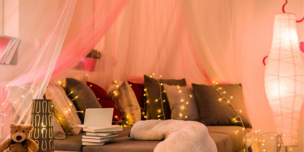 luxury kids and teen bedrooms blog banner featuring plump cushions on grey bedding beside wooden drawers, pink voile curtains and fairy lights