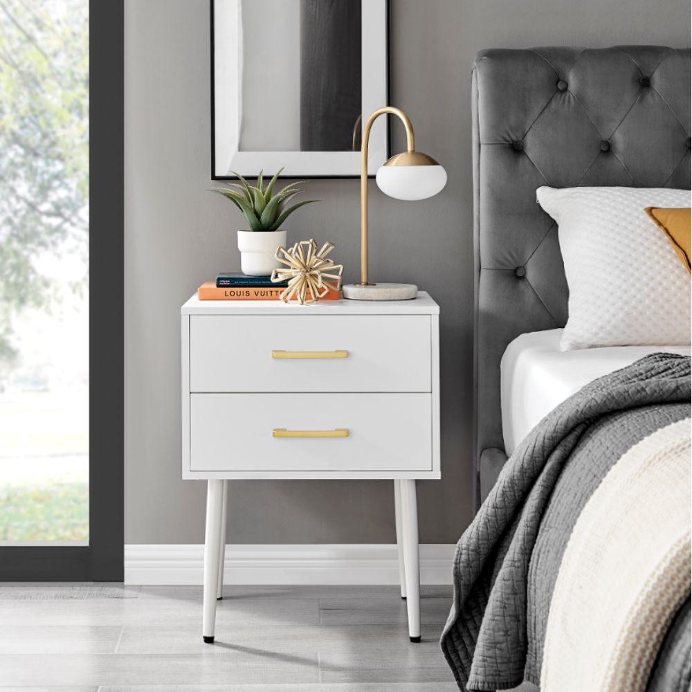 modern minimalist Scandi-inspired bedside table with 2 drawers and tapered legs. In white, with 2 gold bar handles. modern lamp, 2 books and a small succulent are placed on bedside tabletop. 