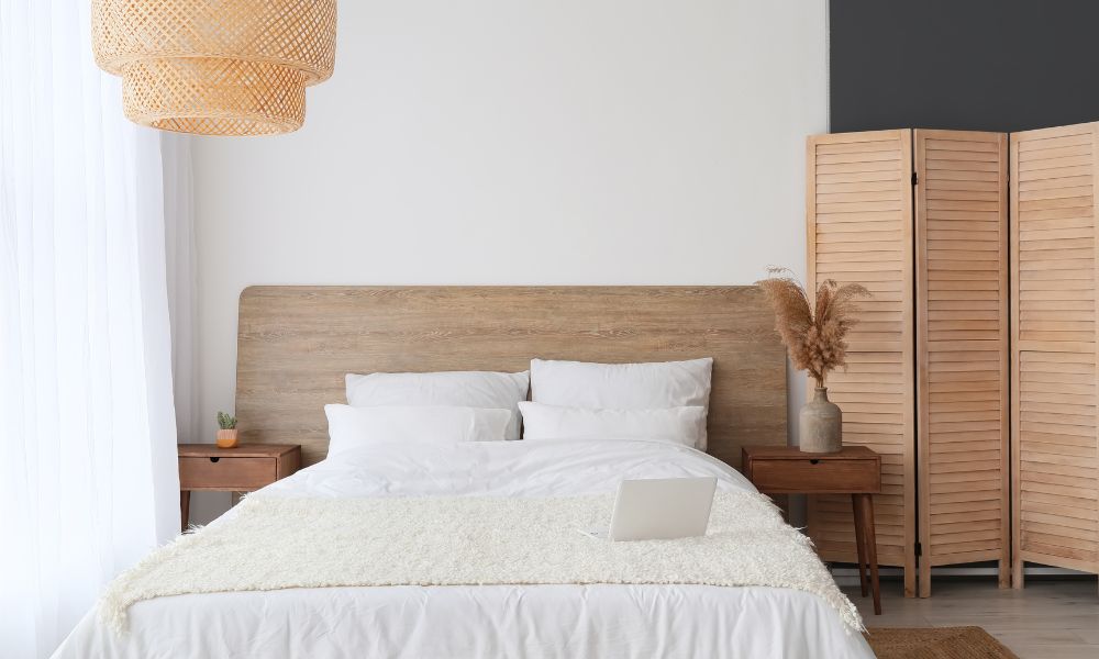 bright airy bedroom with double bed that has large wooden headboard which extends either side of bed behind 2 wooden bedside tables. There is a wooden folding sceen to the right of the image. The bed linens are white and cream. A wicker rattan light fixing hangs off centre, to the left of the photo.