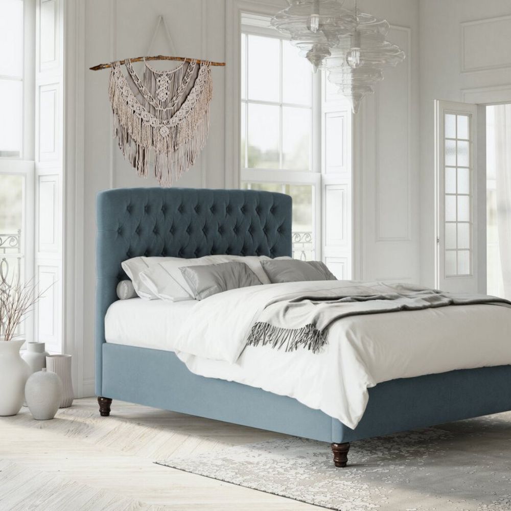 pale blue fabric uphostered statement bed with tall heaboard. Chesterfield style button tufting to the headboards, room is white with pale bed linens. Bed is positioned between 2 windows and at an angle to the door. A macrame wall hanging is hung above the bed. 