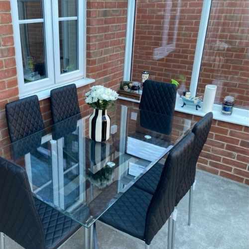 conservatory dining room - rectangular glass dining table with chrome nested legs in a starburst design, with 6 black faux leather modern dining chairs with tall back and chrome legs. In a red brick conservatory with grey concrete floor and white window frames. A black and white vase of white flowers sits on the tabletop.