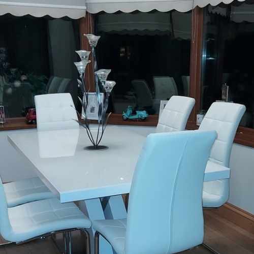 White high gloss dining table with X shaped legs and chrome accents, with 6 white faux leather dining chairs on cantilever legs, in modern conservaotry with brown window frames and white plater walls, and wood flooring. A sculpture of flowers in metal and glass sits on the dining table.