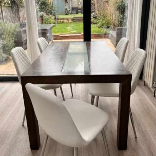 Light warm wood floor in front of wide sliding patio doors with white curtains. Rustic dark brown dining table in front of doors, with 6 modern white faux leather dining chairs all with silver tapered legs. 