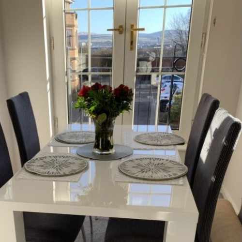 Bright and sunny conservatory with french doors. A white high gloss dining table with simple design and 4 tall-backed dining chairs sit in front of the doors, with placemats and a vase of red roses.