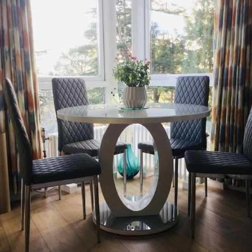 large UPVC windows with patterned curtains and warm wood flooring. Round High gloss dining table with structural halo O shaped plinth leg and grey glass top. 4 grey faux leather tall back dining chairs. Pot plant in centre of table.