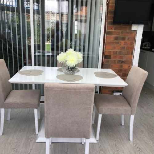 red brick conservatory dining area with tall windows and venetian blinds. White high gloss dining table with 2 column plinth legs in front of windows, with 4 fabric upholstered chairs, placemats and glass vase with cream flowers. Pale wood floor. 