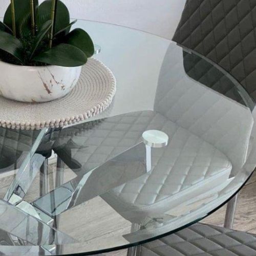 Best Space Saving Dining Tables And Chairs (Perfect For Small Rooms) blog - image shows round clear glass table with chrome legs in nested starburst deisng, with orchid on tabletop and grey faux leather dining chairs pushed up to table.