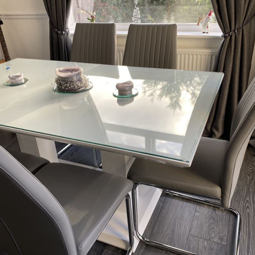 white high gloss dining table from Furniturebox UK, with clear glass table top protector placed on top of it. The table and 6 faux leather chairs are placed in front of a sunny window. 