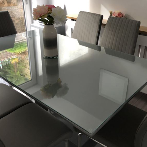 dining table in sunny dining room with 6 grey faux leather dining chairs. The table has a glass table top protector on it. The french windows and garden are reflected in the table top.