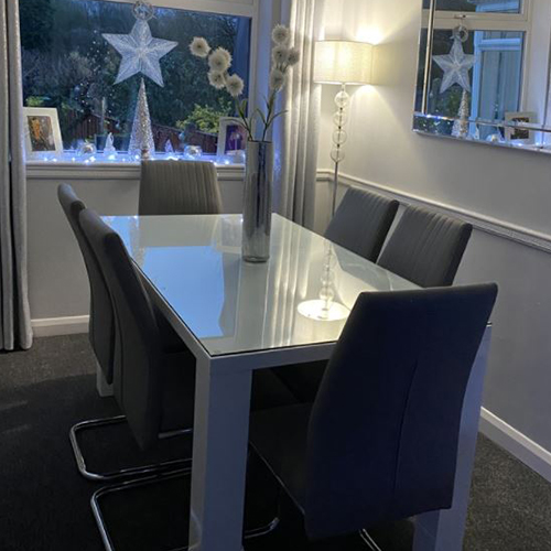 White high gloss Pivero dining table from Furniturebox UK with 6 grey faux leather dining chairs, in a bright white dining room. A glass table top protector is shown placed on top of the table.