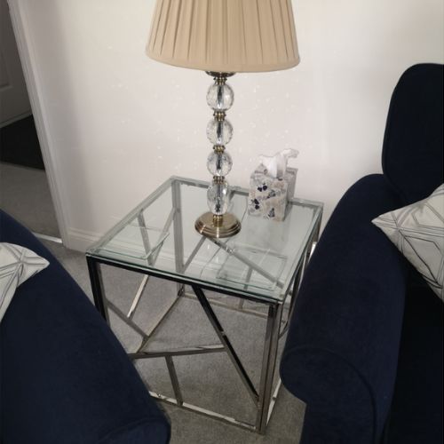 corenr space between two sofas. square cube side table with clear glass top and silver chrome base featuring geometric abstract shapes. Tall table lamp on top,