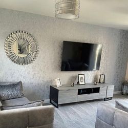 grey and silver living room with abstract silver-flecked wall paper and grey velvet sofas, grey wood TV stand and a round wall mirror in mirrored starburst frame