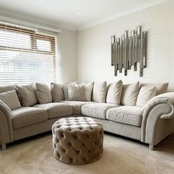 beige living room with cream beige corner sofa, velvet foot stool and abstract modern wall mirror