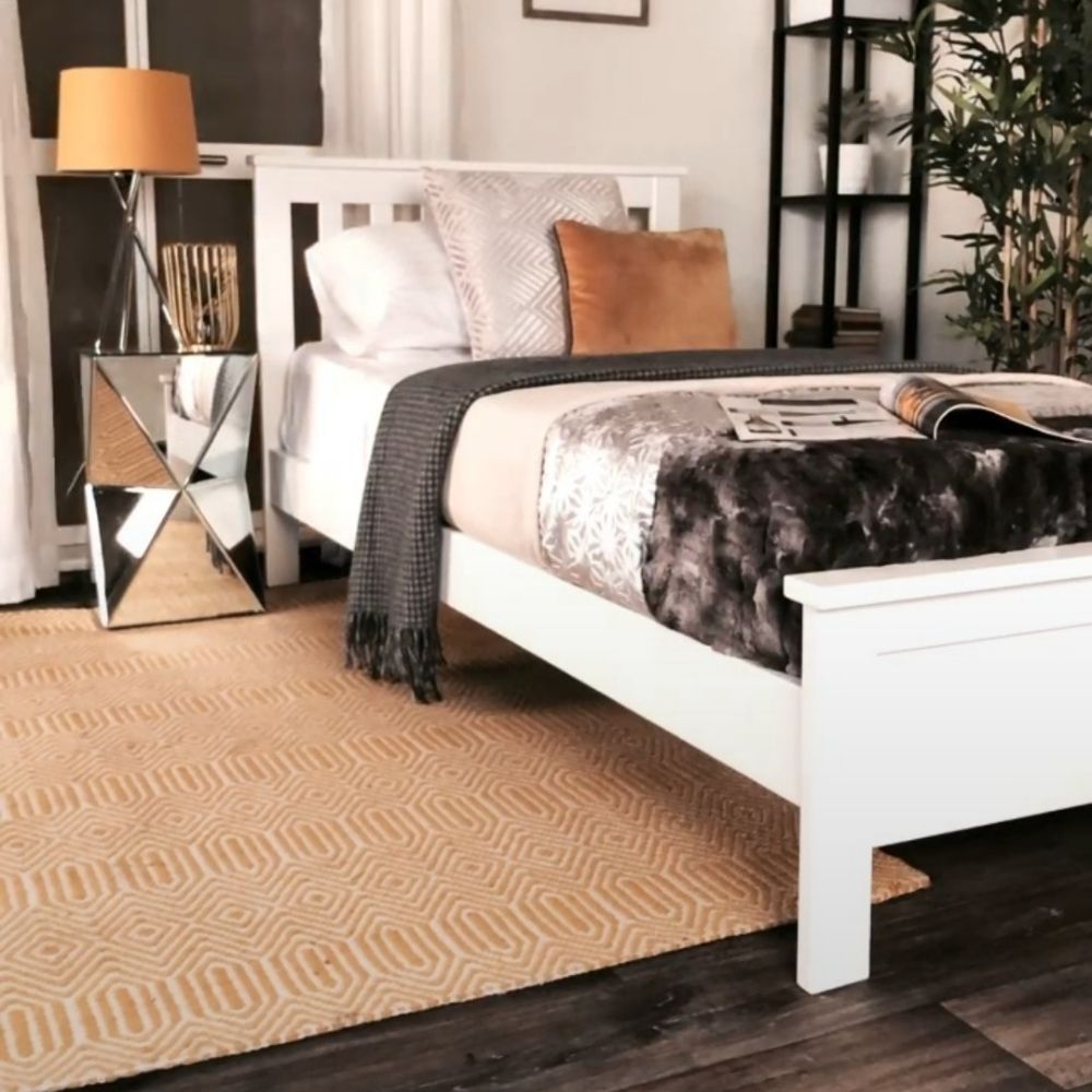 glam bedroom featuring white solid pin bed, white walls, gold rug, mirrored geometric bedside table, chrome lamp with gold shade, gold accessories, black minimalist shelving in corner with books and potplants, grey and silver linens on bed with open magazine. 