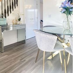 pale wood floor with white walls, round glass table with gold nested starburst legs, 4 grey velvet chairs with gold tapered legs, mirrored console in background, vase of blue lilies on table