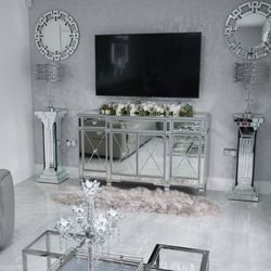 modern living room with grey shiny tiled floor and lots of chrome and mirrored funrniture, with chandelier style candle stick on coffee table, 2 mirrored column plinths either side of mirrored TV stant, and 2 round wall mirrors with crenalated zig zag pattern to frame. 