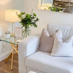 bright but cosy living room featuring pale white grey sofa and white cushions, with square glass end side table that has nested starburt gold metal legs, a cream gold and glass lamo, and white and gold candles, and a vase of white roses. 