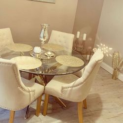 Modern round glass dining table with gold metal legs in a nested starburst design, and 4 white fabric chairs with chesterfield style button details and wooden legs. Gold accessories and candles decorate the table and floor
