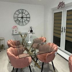 bright dining room with rectangular glass dining table with gold metal nested starburst legs and 4 pink velvet chairs with scalloped backs and tapered legs. A glass console matching the dining table is in the rear. 