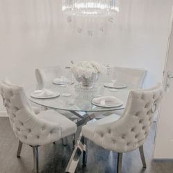 round glass dining table with nested starburst legs and 4 grey velvet knockerback chairs, set with plates and chargers, and a bowl of white roses in centre, with chandelier style light fitting hanging above