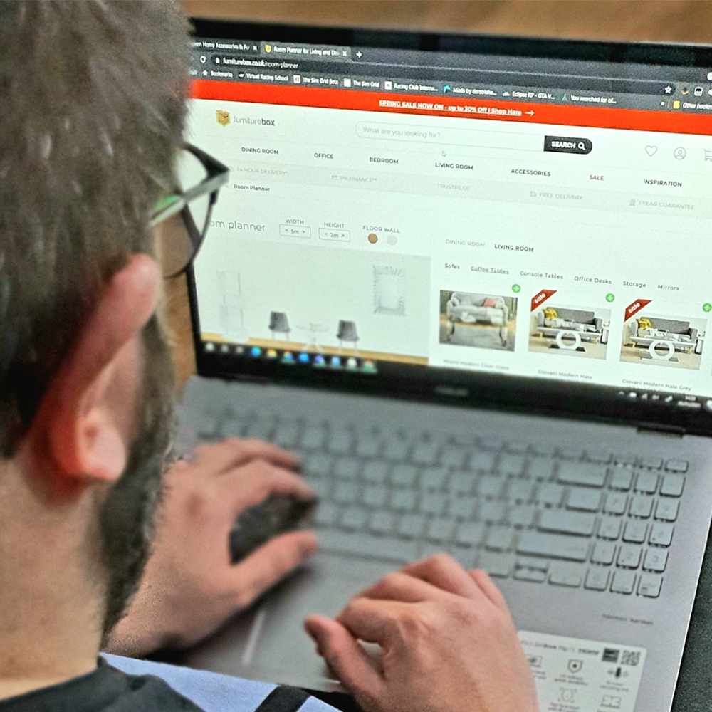 photo over a young bearded man's shoulder as he looks at a laptop sreen featuring the Furniturebox UK Room Planner page