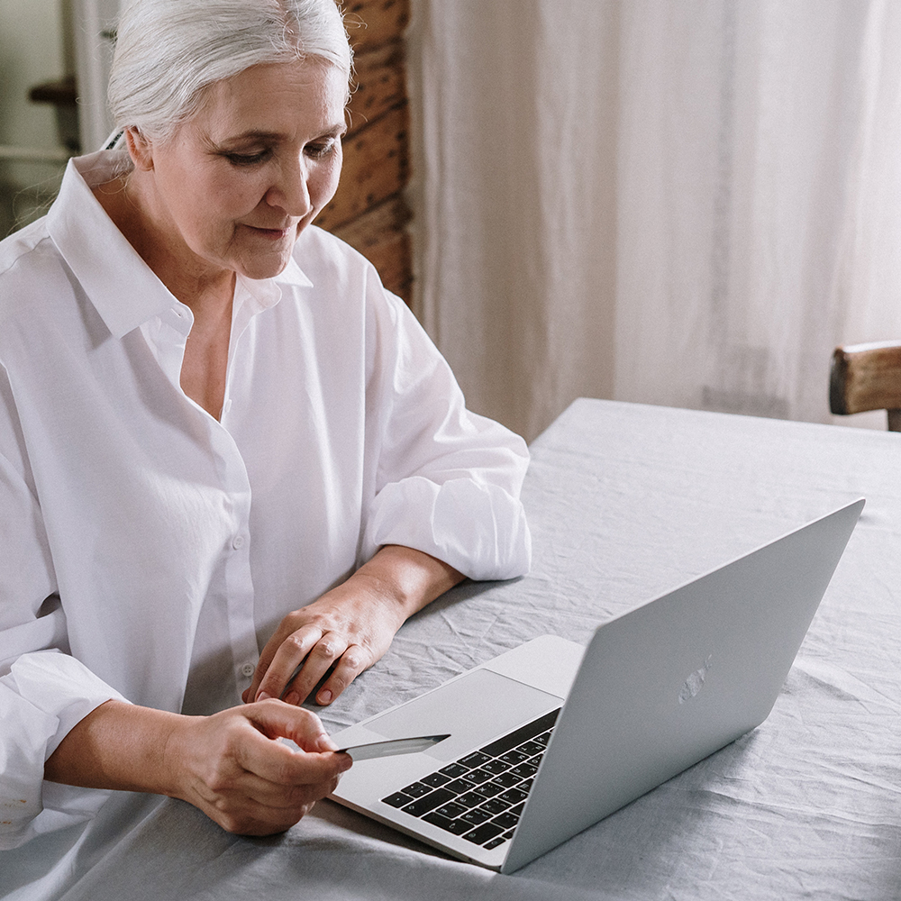 image of older light-skinned woman with white hair  sat at a table looking at her laptop. She is holding a credit card in one hand and smiles.