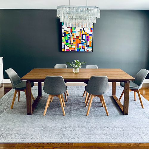 a simple wooden trestle table with 6 bucket seats, all placed on a pale grey rug. the walls are dark grey, with a colourful abstract art print on the wall and modern hanging crystal effect light fixing.