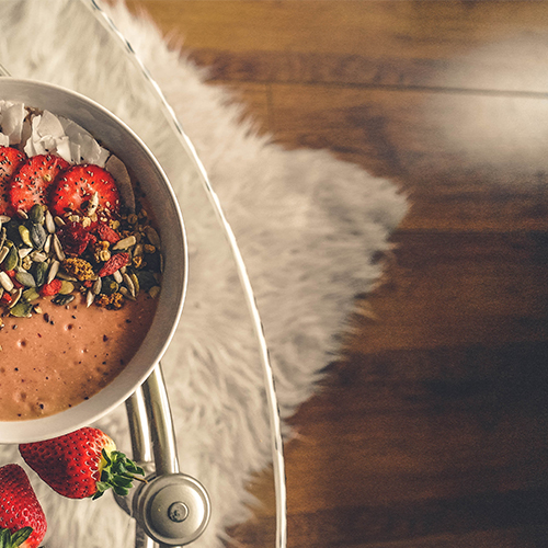 detail shot of a bowl of cereal and fruit on top of a round clear glass dining table. A thick, fluffy shaggy rug can be seen beneath, through the glass tabletop.