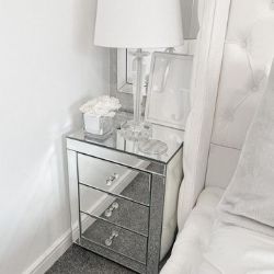 glam reflective 3 drawer bedside table in mirrored surface, with round cut glass drawer knobs, beside white bed, accessorised with white roses, glass and white lamp, and mirror. 