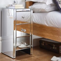 narrow 3 drawer bedside table with round cut glass drawer knobs, beside wood-framed bed in white room. 