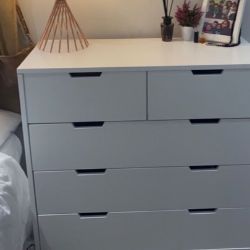 white modern low chest of drawers wit 4 tiers, beside a bed, acting as a bedside table with additional storage. 