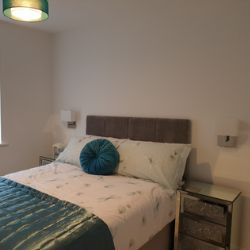 modern bedroom with mirrored crushed glass diamond effect bedside tables that have 3 drawers each, either side of double bed with grey headboard. Lamps are fitted to wall above bedside tables. 