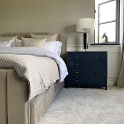 luxury modern bedroom featuring beige velvet upholstered bed from Furnitrebix UK and cream / gold linens, with black chest of drawers bedside bed with lamp.