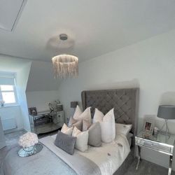 modern grey bedroom with large grey statement bed with chesterfield style headboard, and mirrored & chrome bedside drawers with curved legs. Bed is covered in plump cushions, modern chandelier light fixing. Glam maximalist room