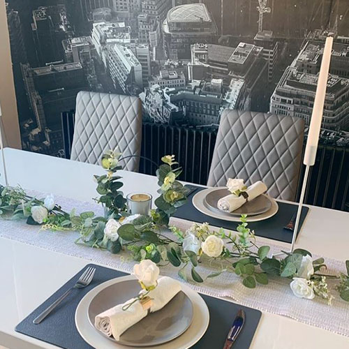 white table with grey faux leather chairs. long table garland of white roses and pael green leaves, and white taper candels. White charger plates, grey dinner plates, white napkins with gold napkin rings and white roses on top.