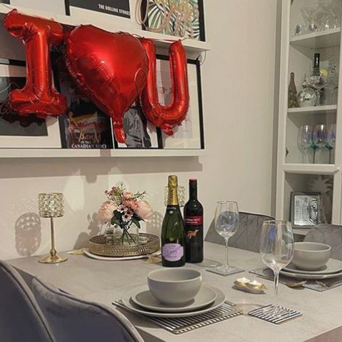 Grey dining table, grey velvet chairs, set with white crockery, bottle of white and prosecco, pink flowers, gold candle holders, and red balloons I "heart" U.