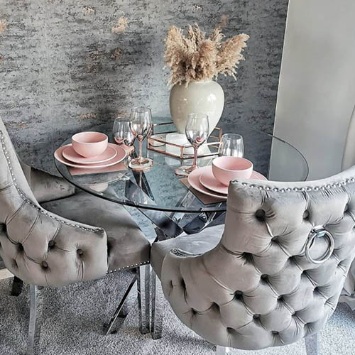 round glass dining table wtih 2 grey velvet chairs. Table dressed with pink crockery and rose gold accessories. Vase of pampas grass. 