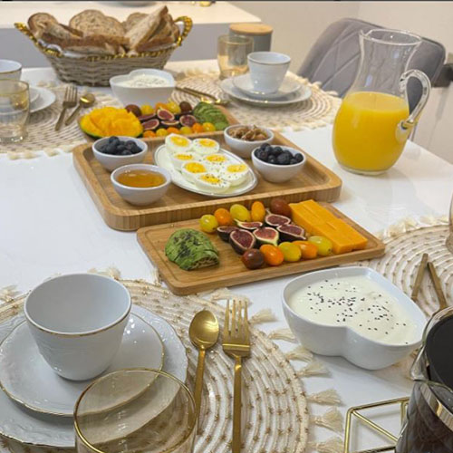 table set for breakfast with jug of range juice, lots of cut fruite, cheeses, eggs, brea, coffee, and white/gold crockery. White table, grey velvet dining chairs.