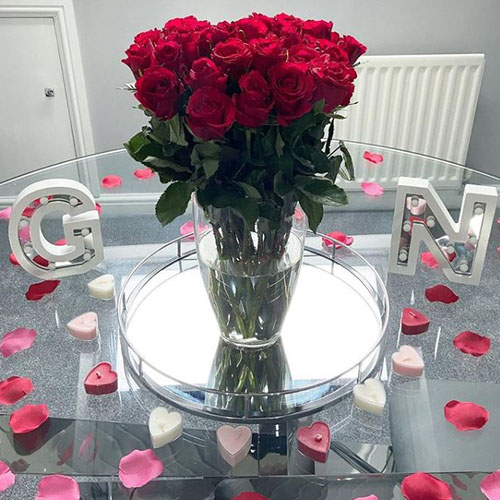 round glass dining table with silver reflective tray, glass vase of red roses, table is scattered with pink and red petals and heart shaped tea-lights.