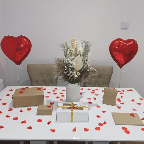 white high-gloss dining table with white ceramic vase, dried flowers and gresses rrnageknet, 2 beige chairs. Table is scattered with red petals, and gifts in borwn and white paper, with 2 heart balloons.