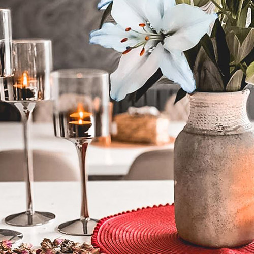 close up of white gloss table wth red placemat, stone vase, white lillies, glass cangle holders and rose buds on table. 