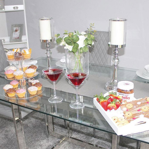 rectangular glass dining table with grey faux leather chairs, with tray of cakes, strawberries and chocolate sprea, cake stand with cupcakes and 2 glasses of red drink in martini glasses. white pillar cnadles and white roses on table. 