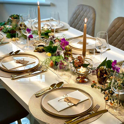 white gloss table with 2 taper candles and long table scpae featuring gold ornaments and flowers in deep purple-red tones with greenery. Table settings are gold.