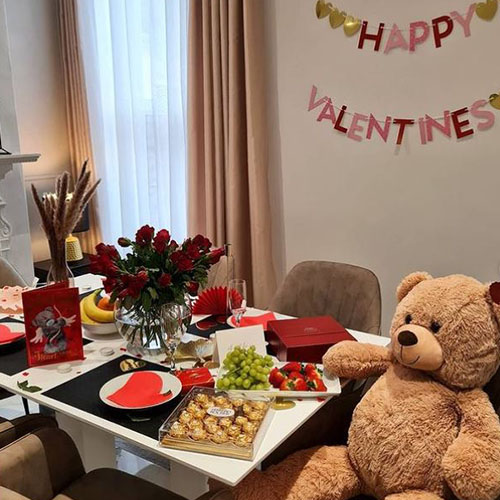 white table, beige chairs, vase of roses, table is dressed for Valentines day with heart napkins, strawberries and grapes, cards and boxes of chocolates, a teddy bear, and banner on wall reading Happy Valentines"