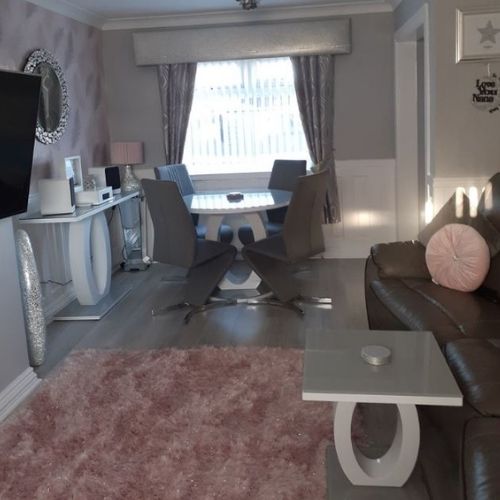 modern living room featuring matching grey glass and high gloss furniture - console table, round dining table and square side table. All tables feature halo structural plinth leg. Pink rug on floor and pink accents to accessories. 
