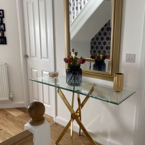 glass and gold metal console table on a landing at top of stiars, beneath rectangular gold mirror