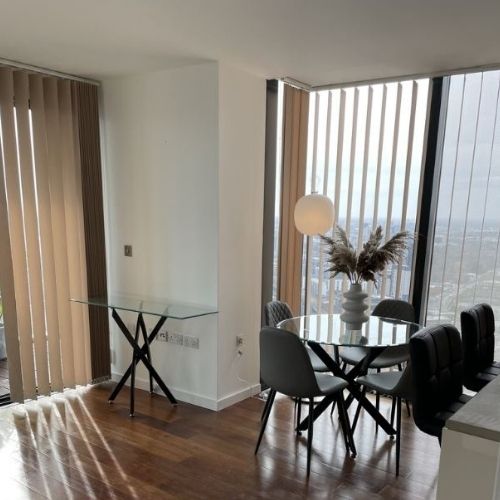 Modern flat / apartment with round glass dining table with black metal nested starburst legs and rectangular glass console table with legs that match dining table. Tall windows and blinds behind wth urban city view beyond. 