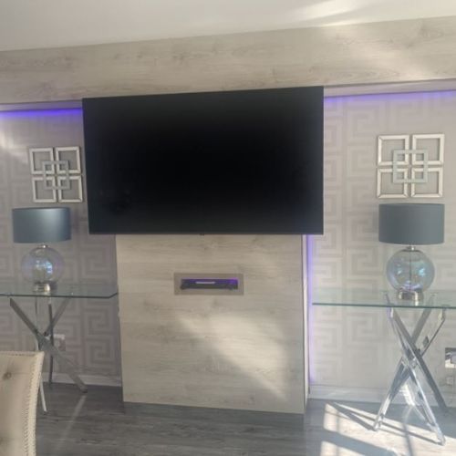 large wall-mounted TV with alcoves either side that have grey and silver key design pattern wallpaper. matching glass and chrome console tables, each with glass and silver lamps, are in the alcoves.