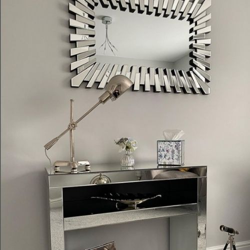 mirrored console table with large rectangular wall mirror hung horizontally above it. mirror has reflective mirrored frame. Console table has ornaments and a silver angle-poise lamp. 