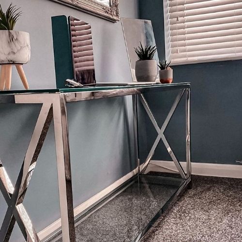 glass and chrome console table with X design to either end of leg frame, in a corner of a room with dark grey walls. A laptop is on the table top.